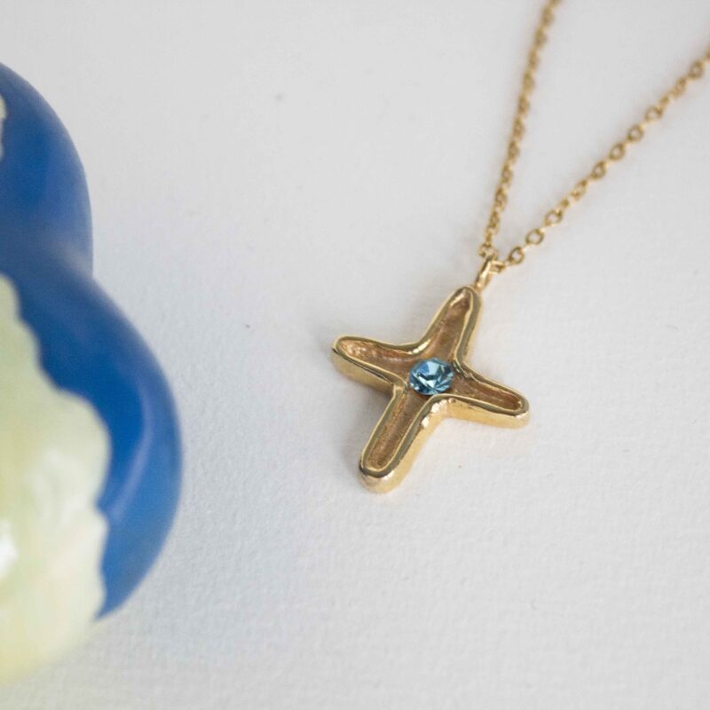 Discover the elegance of the Maria’s Cross necklace with light blue stone, a piece that blends timeless symbolism with artisanal craftsmanship. Handmade from high-quality brass and plated with radiant 24k gold, this cross pendant carries a soft light blue gemstone at its center, symbolizing serenity, hope, and inner peace.Ανακαλύψτε την κομψότητα του κολιέ Maria’s Cross με γαλάζια πέτρα, ένα κόσμημα που συνδυάζει διαχρονικό συμβολισμό με χειροποίητη τέχνη. Κατασκευασμένο στο χέρι από ορείχαλκο υψηλής ποιότητας και επιχρυσωμένο με 24 καράτια χρυσό, το μενταγιόν σε σχήμα σταυρού φιλοξενεί μια γαλάζια πέτρα στο κέντρο του, που συμβολίζει τη γαλήνη, την ελπίδα και την εσωτερική αρμονία.