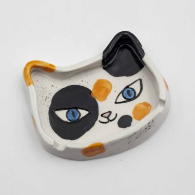 A handmade ceramic calico cat ashtray, featuring a tri-color cat design with orange, black, and white patches, perfect for holding ash and cigarettes. Ένα χειροποίητο κεραμικό τασάκι γάτας κάλικο, με σχέδιο τριχρωμίας γάτας με πορτοκαλί, μαύρες και λευκές περιοχές, ιδανικό για την αποθήκευση στάχτης και τσιγάρων.