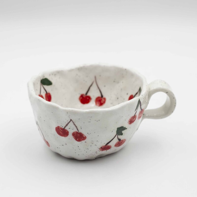 Close-up of a ceramic handmade mug with cherry motifs, featuring vibrant red cherries and green leaves on a white background. Κοντινή λήψη ενός κεραμικού χειροποίητου κύπελου με μοτίβα κερασιών, με έντονα κόκκινα κεράσια και πράσινα φύλλα σε λευκό φόντο.