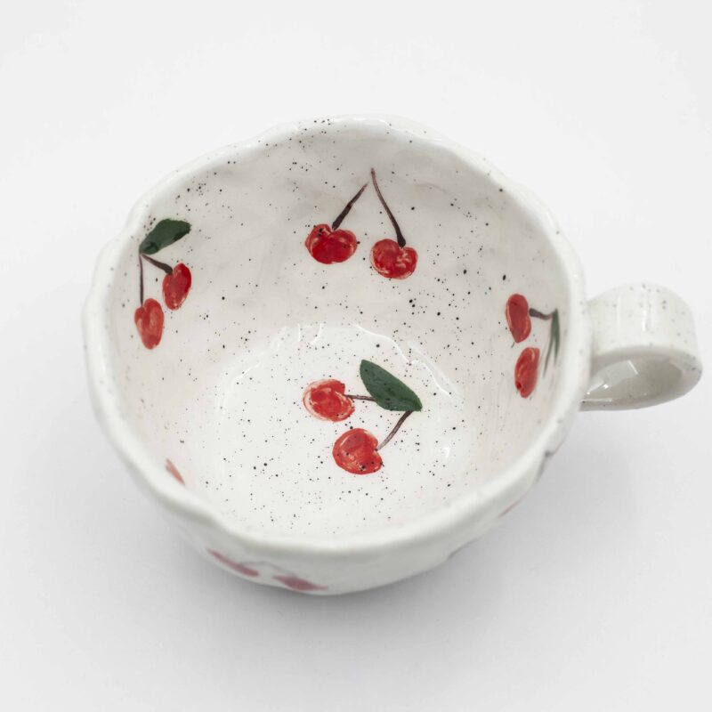 Close-up of a ceramic handmade mug with cherry motifs, featuring vibrant red cherries and green leaves on a white background. Κοντινή λήψη ενός κεραμικού χειροποίητου κύπελου με μοτίβα κερασιών, με έντονα κόκκινα κεράσια και πράσινα φύλλα σε λευκό φόντο.