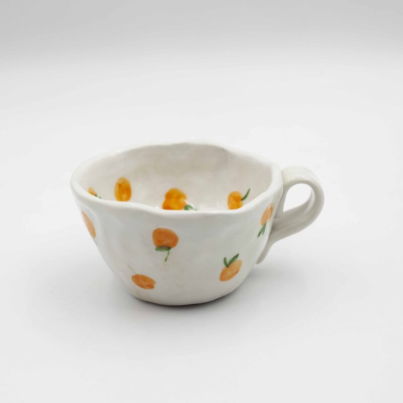 Close-up of a ceramic handmade mug with tangerine motifs, showcasing bright orange tangerines and green leaves on a white background. Κοντινή λήψη μιας χειροποίητης κεραμικής κούπας με μοτίβα μανταρινιών, που επιδεικνύει φωτεινά πορτοκαλί μανταρίνια και πράσινα φύλλα σε λευκό φόντο.