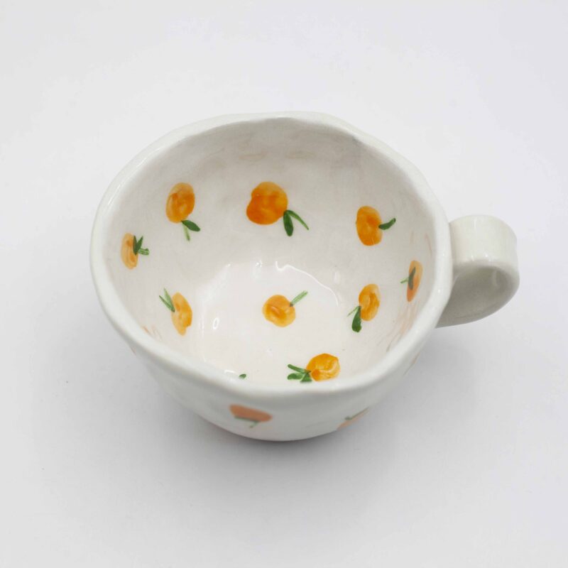 Close-up of a ceramic handmade mug with tangerine motifs, showcasing bright orange tangerines and green leaves on a white background. Κοντινή λήψη μιας χειροποίητης κεραμικής κούπας με μοτίβα μανταρινιών, που επιδεικνύει φωτεινά πορτοκαλί μανταρίνια και πράσινα φύλλα σε λευκό φόντο.