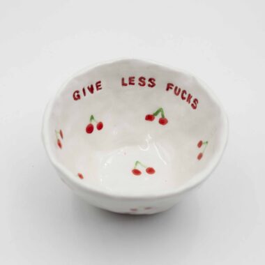 Close-up of a ceramic handmade bowl with cherry motifs and the quote 'Give less fucks' written in stylish typography. Κοντινή λήψη ενός κεραμικού χειροποίητου μπολ με μοτίβα κερασιών και τη φράση 'Give less fucks' γραμμένη με στιλάτη τυπογραφία.