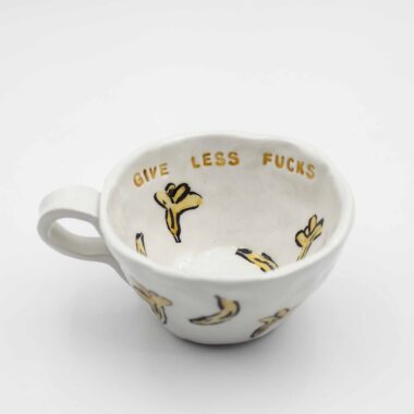 Close-up of a ceramic handmade mug with banana motifs and the quote 'Give less fucks' written in stylish typography. Κοντινή λήψη μιας χειροποίητης κεραμικής κούπας με μοτίβα μπανανών και τη φράση 'Give less fucks' γραμμένη με στιλάτη τυπογραφία.