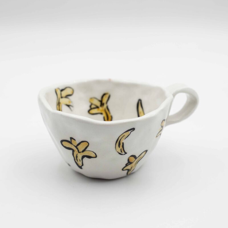 Close-up of a ceramic handmade mug with banana motifs and the quote 'Give less fucks' written in stylish typography. Κοντινή λήψη μιας χειροποίητης κεραμικής κούπας με μοτίβα μπανανών και τη φράση 'Give less fucks' γραμμένη με στιλάτη τυπογραφία.