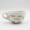 Close-up of a ceramic handmade mug with lemons motifs and the quote 'Give less fucks' written in stylish typography. Κοντινή λήψη μιας χειροποίητης κεραμικής κούπας με μοτίβα λεμονιών και τη φράση 'Give less fucks' γραμμένη με στιλάτη τυπογραφία.