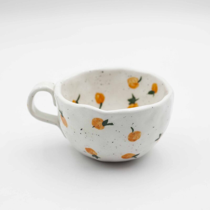 Close-up of a ceramic handmade mug with orange motifs, showcasing vibrant oranges and green leaves on a white background. Κοντινή λήψη ενός κεραμικού χειροποίητου κύπελου με μοτίβα πορτοκαλιών, επιδεικνύοντας ζωηρά πορτοκάλια και πράσινα φύλλα σε λευκό φόντο.