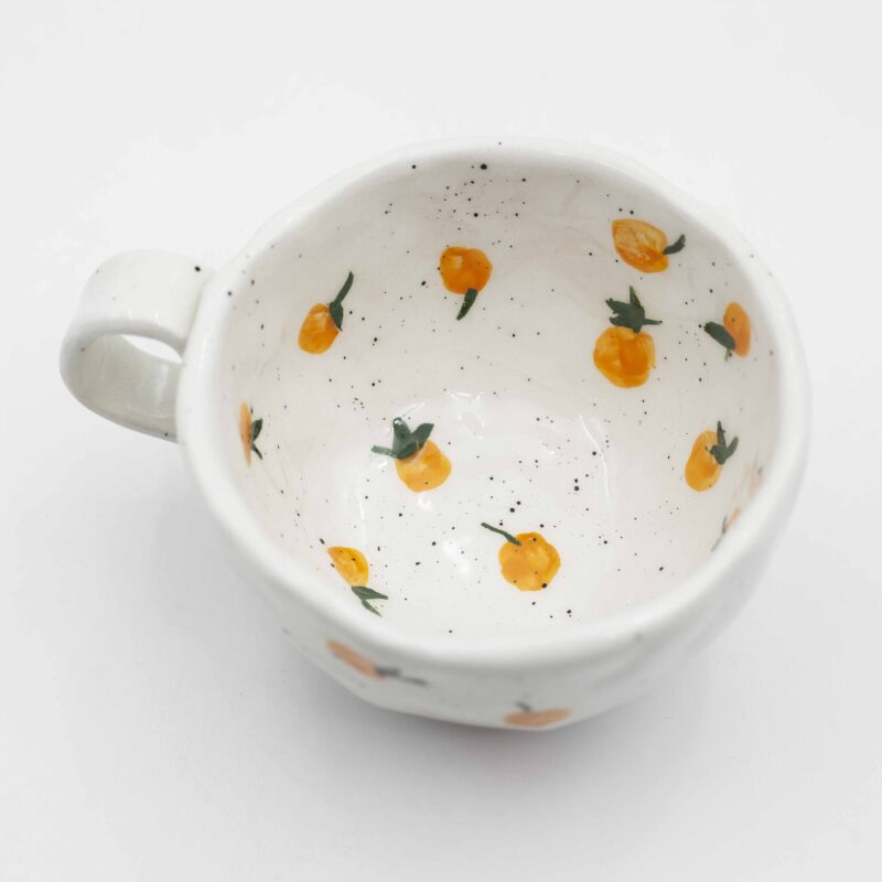 Close-up of a ceramic handmade mug with orange motifs, showcasing vibrant oranges and green leaves on a white background. Κοντινή λήψη ενός κεραμικού χειροποίητου κύπελου με μοτίβα πορτοκαλιών, επιδεικνύοντας ζωηρά πορτοκάλια και πράσινα φύλλα σε λευκό φόντο.