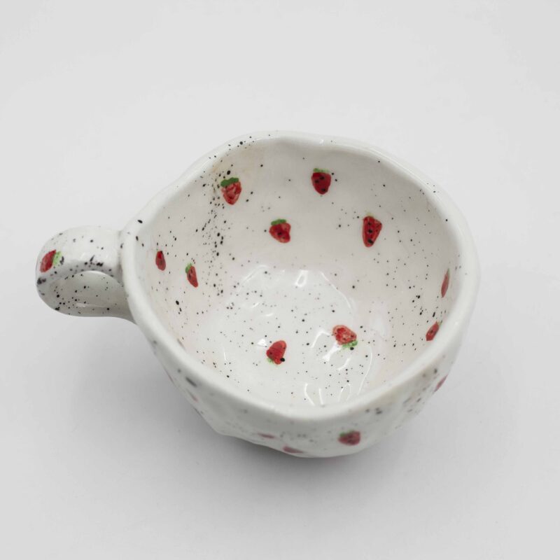 Close-up of a ceramic handmade mug with strawberry motifs, showcasing juicy red strawberries and green leaves on a white background. Κοντινή λήψη ενός κεραμικού χειροποίητου κύπελλου με μοτίβα φράουλας, επιδεικνύοντας χυμώδεις κόκκινες φράουλες και πράσινα φύλλα σε λευκό φόντο.