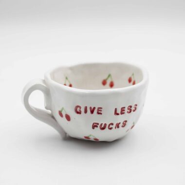 Close-up of a ceramic handmade mug with cherry motifs and the quote 'Give less fucks' written in stylish typography. Κοντινή λήψη μιας χειροποίητης κεραμικής κούπας με μοτίβα κερασιών και τη φράση 'Give less fucks' γραμμένη με στιλάτη τυπογραφία.