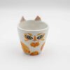 Close-up of a ceramic handmade cat orange mug, featuring a playful cat design with orange fur and green eyes on a white background. Κοντινή λήψη ενός κεραμικού χειροποίητου κύπελλου με μοτίβο παιχνιδιάρικης γάτας με πορτοκαλί τρίχα και πράσινα μάτια σε λευκό φόντο.