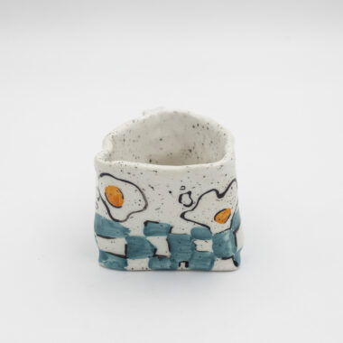 Μια χειροποίητη κεραμική κούπα με αυγά και γαλάζιο πάτωμα σκακιέρα που δημιουργεί μια σουρεαλιστική αίσθηση. English: A handmade ceramic mug adorned with eggs, creating a surrealistic sensation.