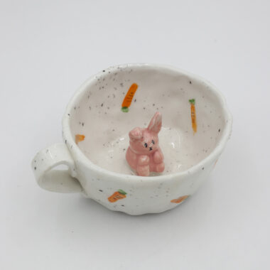 Μια χειροποίητη κεραμική κούπα με σχέδια καρότων πάνω της και μέσα σε αυτήν ένα 3D κουνελάκι. A handmade ceramic mug with carrot drawings on it, and inside it, a 3D bunny.