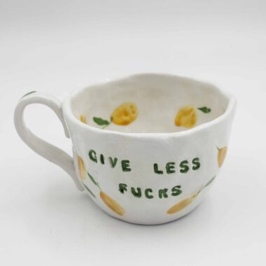 Close-up of a ceramic handmade mug with lemon motifs and the quote 'Give less fucks' written in stylish typography. Κοντινή λήψη μιας χειροποίητης κεραμικής κούπας με μοτίβα λεμονιών και τη φράση 'Give less fucks' γραμμένη με στιλάτη τυπογραφία.