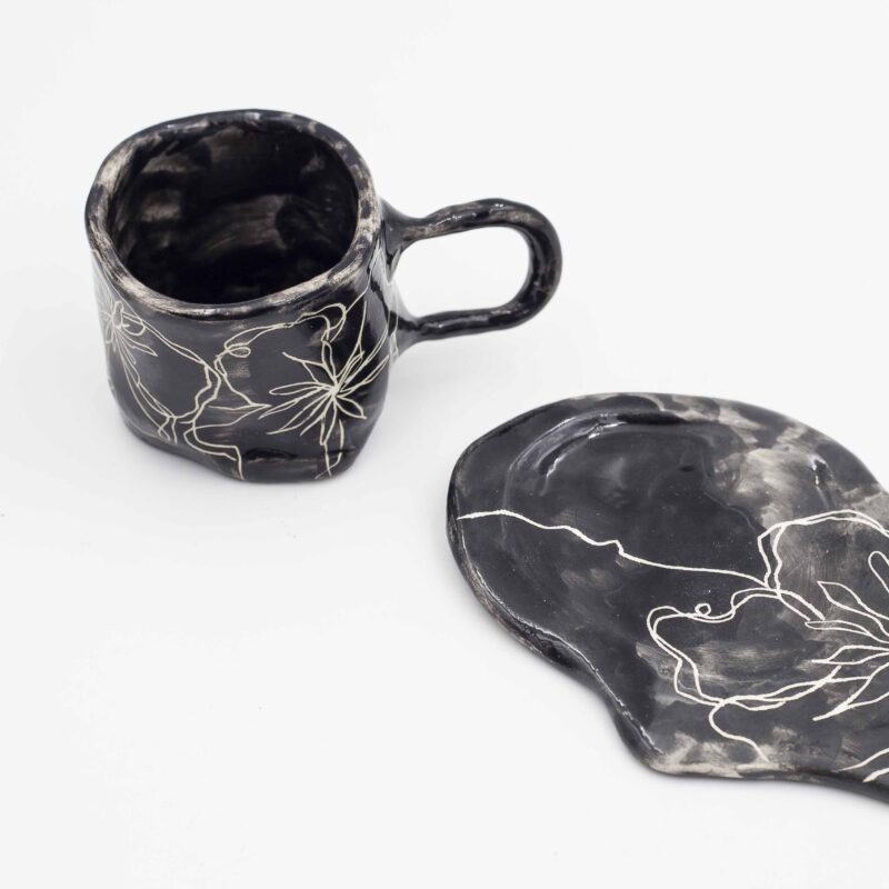 Black ceramic mug with curved floral design, paired with a matching black saucer. Perfect for enjoying your favorite beverage in style. keywords: black mug, floral mug, curved flowers, mug with saucer, ceramic mug Μαύρο φλιτζάνι από κεραμικό με ανάγλυφο floral σχέδιο, μαζί με μαύρο πιατάκι. Ιδανικό για να απολαύσετε το αγαπημένο σας ρόφημα με στυλ. keywords: μαύρο φλιτζάνι, floral φλιτζάνι, ανάγλυφα λουλούδια, φλιτζάνι με πιατάκι, κεραμικό φλιτζάνι