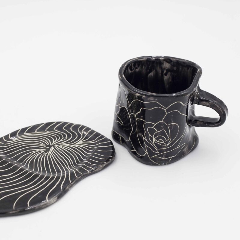 Black ceramic mug with curved floral design, paired with a matching black saucer. Perfect for enjoying your favorite beverage in style. keywords: black mug, floral mug, curved flowers, mug with saucer, ceramic mug Μαύρο φλιτζάνι από κεραμικό με ανάγλυφο floral σχέδιο, μαζί με μαύρο πιατάκι. Ιδανικό για να απολαύσετε το αγαπημένο σας ρόφημα με στυλ. keywords: μαύρο φλιτζάνι, floral φλιτζάνι, ανάγλυφα λουλούδια, φλιτζάνι με πιατάκι, κεραμικό φλιτζάνι