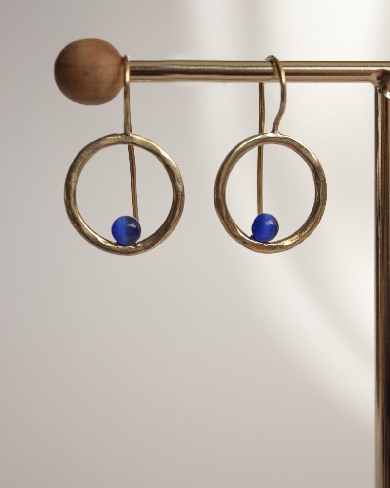 Handmade brass Phaidra circle earrings featuring beautiful blue cat eye stones, crafted by Sunny Designs. These elegant and stylish earrings are perfect for adding a touch of sophistication to any outfit and make a wonderful gift for women. Phaidra earrings, circle earrings, blue cat eye earrings, handmade brass jewelry, Sunny Designs earrings, unique gift, stylish earrings, women’s jewelry, artisan earrings, elegant earrings Χειροποίητα σκουλαρίκια Phaidra σε σχήμα κύκλου από μπρούντζο με όμορφες μπλε πέτρες ματιού της γάτας, φτιαγμένα από τη Sunny Designs. Αυτά τα κομψά και στιλάτα σκουλαρίκια είναι ιδανικά για να προσθέσουν μια αίσθηση κομψότητας σε κάθε ντύσιμο και αποτελούν μια υπέροχη επιλογή δώρου για γυναίκες. σκουλαρίκια Phaidra, κυκλικά σκουλαρίκια, σκουλαρίκια με μπλε μάτι γάτας, χειροποίητο κόσμημα από μπρούντζο, σκουλαρίκια Sunny Designs, μοναδικό δώρο, κομψά σκουλαρίκια, γυναικείο κόσμημα, κοσμήματα τέχνης, κομψά σκουλαρίκια