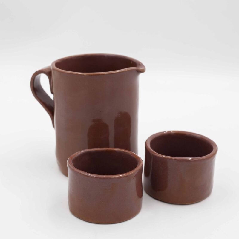 Handmade ceramic jug with two matching cups, crafted from rich red clay and finished with a transparent glaze that beautifully reveals the natural earthy tones. This artisanal set combines functionality with elegance, making it perfect for serving beverages with a rustic, handmade charm. Ideal for adding a touch of natural beauty to your kitchen or table decor. Χειροποίητη κεραμική κανάτα με δύο αντίστοιχες κούπες, κατασκευασμένες από κόκκινο πηλό και καλυμμένες με διάφανο γυαλί που αναδεικνύει τους φυσικούς γήινους τόνους. Αυτό το καλλιτεχνικό σετ συνδυάζει λειτουργικότητα με κομψότητα, ιδανικό για το σερβίρισμα ροφημάτων με μια ρουστίκ, χειροποίητη αίσθηση. Ιδανικό για να προσθέσετε μια φυσική ομορφιά στην κουζίνα ή το τραπέζι σας.