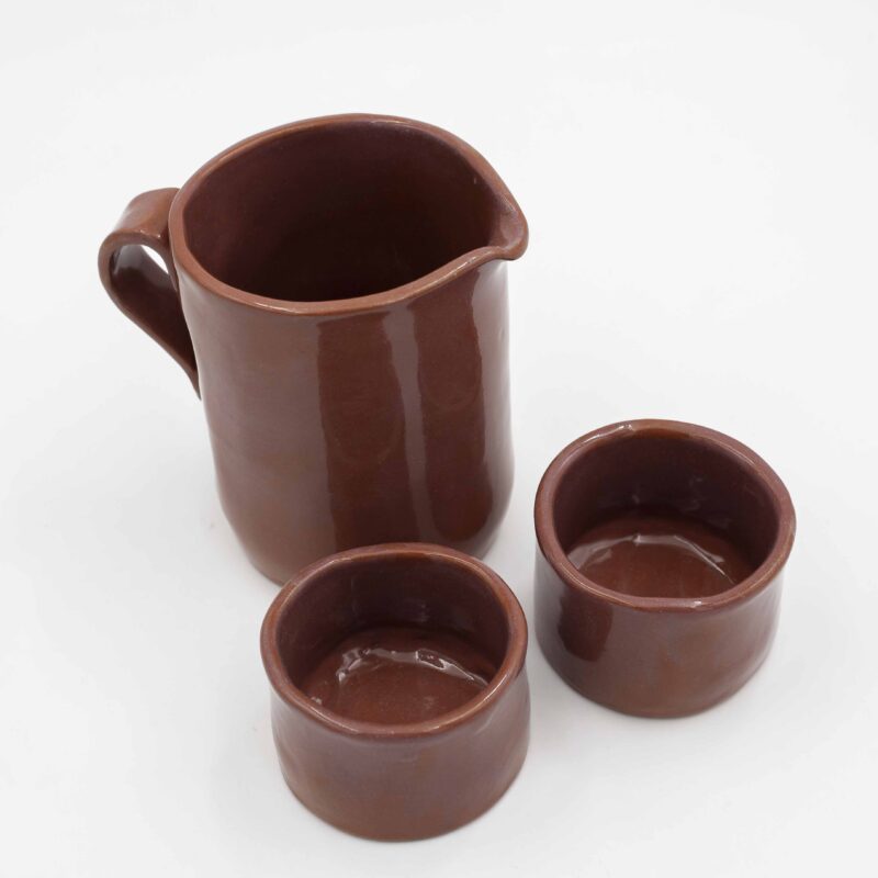 Handmade ceramic jug with two matching cups, crafted from rich red clay and finished with a transparent glaze that beautifully reveals the natural earthy tones. This artisanal set combines functionality with elegance, making it perfect for serving beverages with a rustic, handmade charm. Ideal for adding a touch of natural beauty to your kitchen or table decor. Χειροποίητη κεραμική κανάτα με δύο αντίστοιχες κούπες, κατασκευασμένες από κόκκινο πηλό και καλυμμένες με διάφανο γυαλί που αναδεικνύει τους φυσικούς γήινους τόνους. Αυτό το καλλιτεχνικό σετ συνδυάζει λειτουργικότητα με κομψότητα, ιδανικό για το σερβίρισμα ροφημάτων με μια ρουστίκ, χειροποίητη αίσθηση. Ιδανικό για να προσθέσετε μια φυσική ομορφιά στην κουζίνα ή το τραπέζι σας.