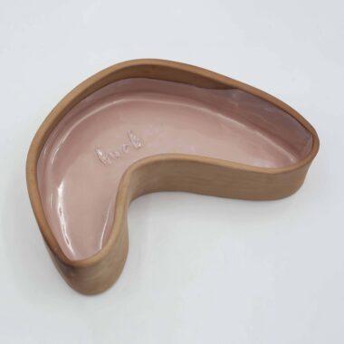 Handmade ceramic boomerang-shaped plate with a unique pink and green glass inscription at the bottom, crafted from red clay. This artistic dish combines a vibrant color palette and organic design, ideal for adding a touch of elegance and originality to any home decor. Χειροποίητο κεραμικό πιάτο σε σχήμα μπούμερανγκ με μοναδική ροζ και πράσινη υάλωση στον πάτο, φτιαγμένο από κόκκινο πηλό. Αυτό το καλλιτεχνικό πιάτο συνδυάζει ζωντανά χρώματα και οργανικό σχέδιο, ιδανικό για να προσθέσει κομψότητα και πρωτοτυπία σε κάθε διακόσμηση σπιτιού.