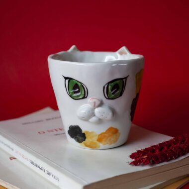 Handmade ceramic mug with a cat face design, available in calico, tuxedo, white, and orange cat variations. Perfect for cat lovers looking to add a cute, personalized touch to their drinkware collection.