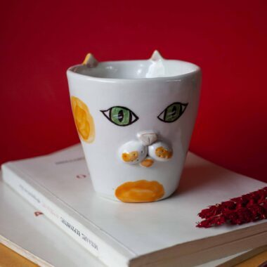 Handmade ceramic mug with a cat face design, available in calico, tuxedo, white, and orange cat variations. Perfect for cat lovers looking to add a cute, personalized touch to their drinkware collection." Greek: "Χειροποίητη κεραμική κούπα με σχέδιο προσώπου γάτας, διαθέσιμη σε εκδόσεις τρίχρωμης, δίχρωμης, λευκής και πορτοκαλί γάτας. Ιδανική για φιλόζωους που θέλουν να προσθέσουν μια χαριτωμένη, προσωπική πινελιά στη συλλογή τους από κούπες.