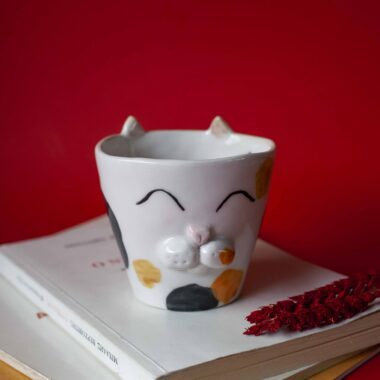 Handmade ceramic mug with a cat face design, available in calico, tuxedo, white, and orange cat variations. Perfect for cat lovers looking to add a cute, personalized touch to their drinkware collection.