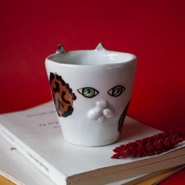 Handmade ceramic mug with a cat face design, available in calico, tuxedo, white, and orange cat variations. Perfect for cat lovers looking to add a cute, personalized touch to their drinkware collection.