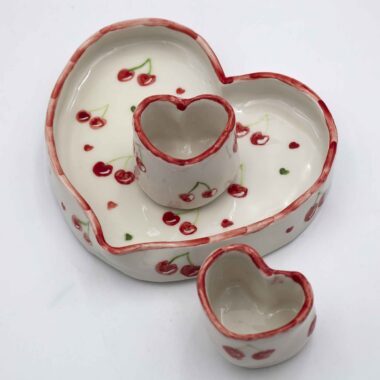 Handmade ceramic heart-shaped set, featuring a large heart plate and two matching heart cups, beautifully hand-painted with cherries. A romantic and charming design, perfect for sharing special moments over coffee or tea. Ideal for Valentine's Day, anniversaries, or as a unique gift for someone you love." Χειροποίητο κεραμικό σετ σε σχήμα καρδιάς, που περιλαμβάνει ένα μεγάλο πιάτο καρδιά και δύο ασορτί κούπες, διακοσμημένα με ζωγραφισμένα στο χέρι κεράσια. Ένα ρομαντικό και γοητευτικό σχέδιο, ιδανικό για να μοιράζεστε ξεχωριστές στιγμές με καφέ ή τσάι. Τέλειο για την Ημέρα του Αγίου Βαλεντίνου, επετείους ή ως μοναδικό δώρο για ένα αγαπημένο σας πρόσωπο.