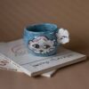 Handmade ceramic mug with a cloud-shaped handle and painted cloud faces featuring shiny eyes in an angelic core aesthetic. Soft blue and pink tones create a dreamy, sweet atmosphere. Each mug is one of a kind — hand-shaped and hand-painted, a small personal piece of art for those who love unique creations. Χειροποίητη κεραμική κούπα με χερούλι σε σχήμα συννεφιού και ζωγραφιστά συννεφάκια με εκφραστικά προσωπάκια και γυαλιστερά μάτια σε angelic core αισθητική. Οι απαλοί τόνοι του γαλάζιου και του ροζ προσδίδουν μια ονειρική, γλυκιά αίσθηση. Κάθε κούπα είναι μοναδική, πλασμένη και ζωγραφισμένη στο χέρι, ένα μικρό προσωπικό έργο τέχνης για κάθε λάτρη των ιδιαίτερων αντικειμένων.