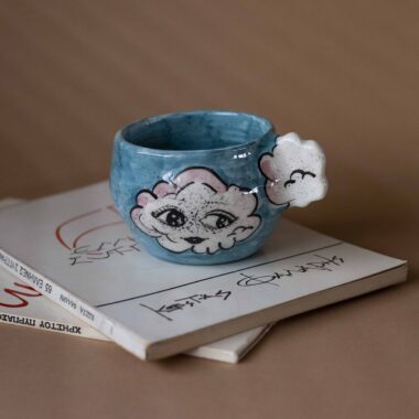 Handmade ceramic mug with a cloud-shaped handle and painted cloud faces featuring shiny eyes in an angelic core aesthetic. Soft blue and pink tones create a dreamy, sweet atmosphere. Each mug is one of a kind — hand-shaped and hand-painted, a small personal piece of art for those who love unique creations. Χειροποίητη κεραμική κούπα με χερούλι σε σχήμα συννεφιού και ζωγραφιστά συννεφάκια με εκφραστικά προσωπάκια και γυαλιστερά μάτια σε angelic core αισθητική. Οι απαλοί τόνοι του γαλάζιου και του ροζ προσδίδουν μια ονειρική, γλυκιά αίσθηση. Κάθε κούπα είναι μοναδική, πλασμένη και ζωγραφισμένη στο χέρι, ένα μικρό προσωπικό έργο τέχνης για κάθε λάτρη των ιδιαίτερων αντικειμένων.