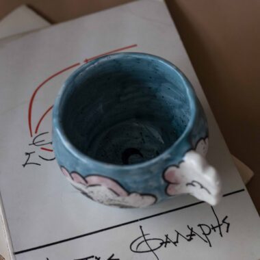 Handmade ceramic mug with a cloud-shaped handle and painted cloud faces featuring shiny eyes in an angelic core aesthetic. Soft blue and pink tones create a dreamy, sweet atmosphere. Each mug is one of a kind — hand-shaped and hand-painted, a small personal piece of art for those who love unique creations. Χειροποίητη κεραμική κούπα με χερούλι σε σχήμα συννεφιού και ζωγραφιστά συννεφάκια με εκφραστικά προσωπάκια και γυαλιστερά μάτια σε angelic core αισθητική. Οι απαλοί τόνοι του γαλάζιου και του ροζ προσδίδουν μια ονειρική, γλυκιά αίσθηση. Κάθε κούπα είναι μοναδική, πλασμένη και ζωγραφισμένη στο χέρι, ένα μικρό προσωπικό έργο τέχνης για κάθε λάτρη των ιδιαίτερων αντικειμένων.