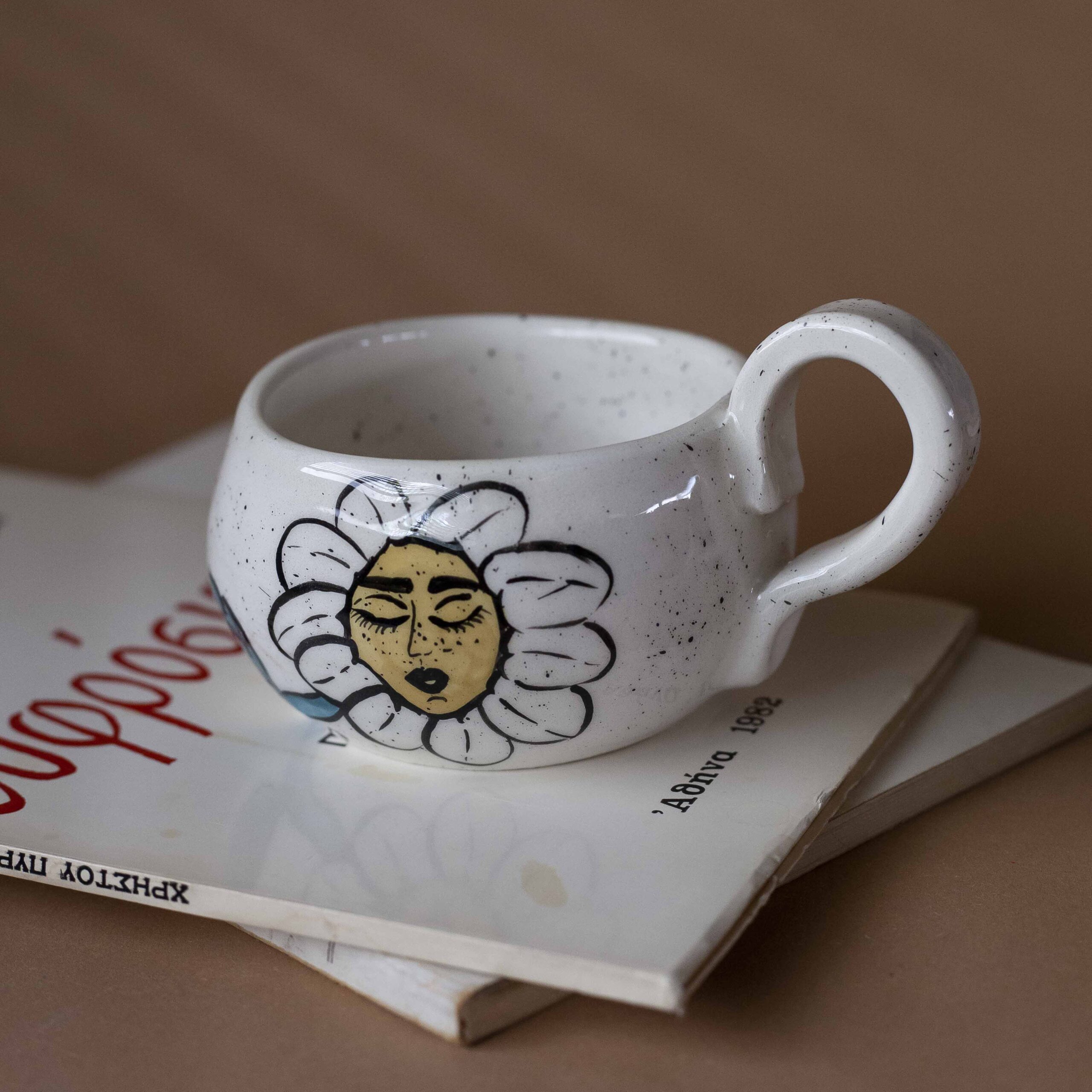 Χειροποίητη κεραμική κούπα διακοσμημένη με ζωγραφιστές μαργαρίτες και πολύχρωμα λουλούδια, όλα με γλυκά προσωπάκια και εκφραστικά μάτια. Πλασμένη και ζωγραφισμένη στο χέρι, κάθε κούπα είναι ένα μοναδικό έργο τέχνης που συνδυάζει τη φρεσκάδα της φύσης με μια παιχνιδιάρικη, ονειρική αισθητική. Ιδανική για όσους αγαπούν τα λουλούδια και τη χειροποίητη δημιουργία. Handmade ceramic mug decorated with painted daisies and colorful flowers, each featuring cute little faces with expressive eyes. Hand-shaped and hand-painted, every mug is a one-of-a-kind piece of art that blends the freshness of nature with a playful, dreamy aesthetic. Perfect for flower lovers and fans of handmade craftsmanship.