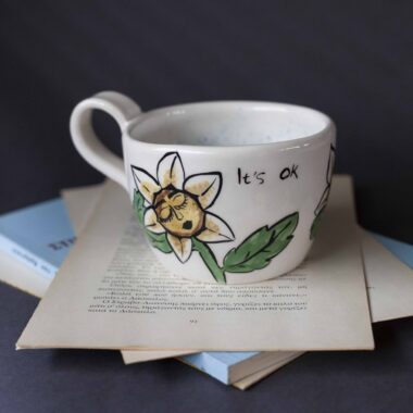 Χειροποίητη κεραμική κούπα διακοσμημένη με ζωγραφιστές μαργαρίτες και πολύχρωμα λουλούδια, όλα με γλυκά προσωπάκια και εκφραστικά μάτια. Πλασμένη και ζωγραφισμένη στο χέρι, κάθε κούπα είναι ένα μοναδικό έργο τέχνης που συνδυάζει τη φρεσκάδα της φύσης με μια παιχνιδιάρικη, ονειρική αισθητική. Ιδανική για όσους αγαπούν τα λουλούδια και τη χειροποίητη δημιουργία. Handmade ceramic mug decorated with painted daisies and colorful flowers, each featuring cute little faces with expressive eyes. Hand-shaped and hand-painted, every mug is a one-of-a-kind piece of art that blends the freshness of nature with a playful, dreamy aesthetic. Perfect for flower lovers and fans of handmade craftsmanship.