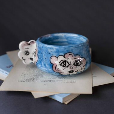 Handmade ceramic mug with a cloud-shaped handle and painted cloud faces featuring shiny eyes in an angelic core aesthetic. Soft blue and pink tones create a dreamy, sweet atmosphere. Each mug is one of a kind — hand-shaped and hand-painted, a small personal piece of art for those who love unique creations. Χειροποίητη κεραμική κούπα με χερούλι σε σχήμα συννεφιού και ζωγραφιστά συννεφάκια με εκφραστικά προσωπάκια και γυαλιστερά μάτια σε angelic core αισθητική. Οι απαλοί τόνοι του γαλάζιου και του ροζ προσδίδουν μια ονειρική, γλυκιά αίσθηση. Κάθε κούπα είναι μοναδική, πλασμένη και ζωγραφισμένη στο χέρι, ένα μικρό προσωπικό έργο τέχνης για κάθε λάτρη των ιδιαίτερων αντικειμένων.