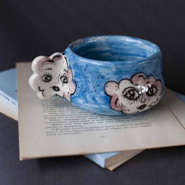 Handmade ceramic mug with a cloud-shaped handle and painted cloud faces featuring shiny eyes in an angelic core aesthetic. Soft blue and pink tones create a dreamy, sweet atmosphere. Each mug is one of a kind — hand-shaped and hand-painted, a small personal piece of art for those who love unique creations. Χειροποίητη κεραμική κούπα με χερούλι σε σχήμα συννεφιού και ζωγραφιστά συννεφάκια με εκφραστικά προσωπάκια και γυαλιστερά μάτια σε angelic core αισθητική. Οι απαλοί τόνοι του γαλάζιου και του ροζ προσδίδουν μια ονειρική, γλυκιά αίσθηση. Κάθε κούπα είναι μοναδική, πλασμένη και ζωγραφισμένη στο χέρι, ένα μικρό προσωπικό έργο τέχνης για κάθε λάτρη των ιδιαίτερων αντικειμένων.