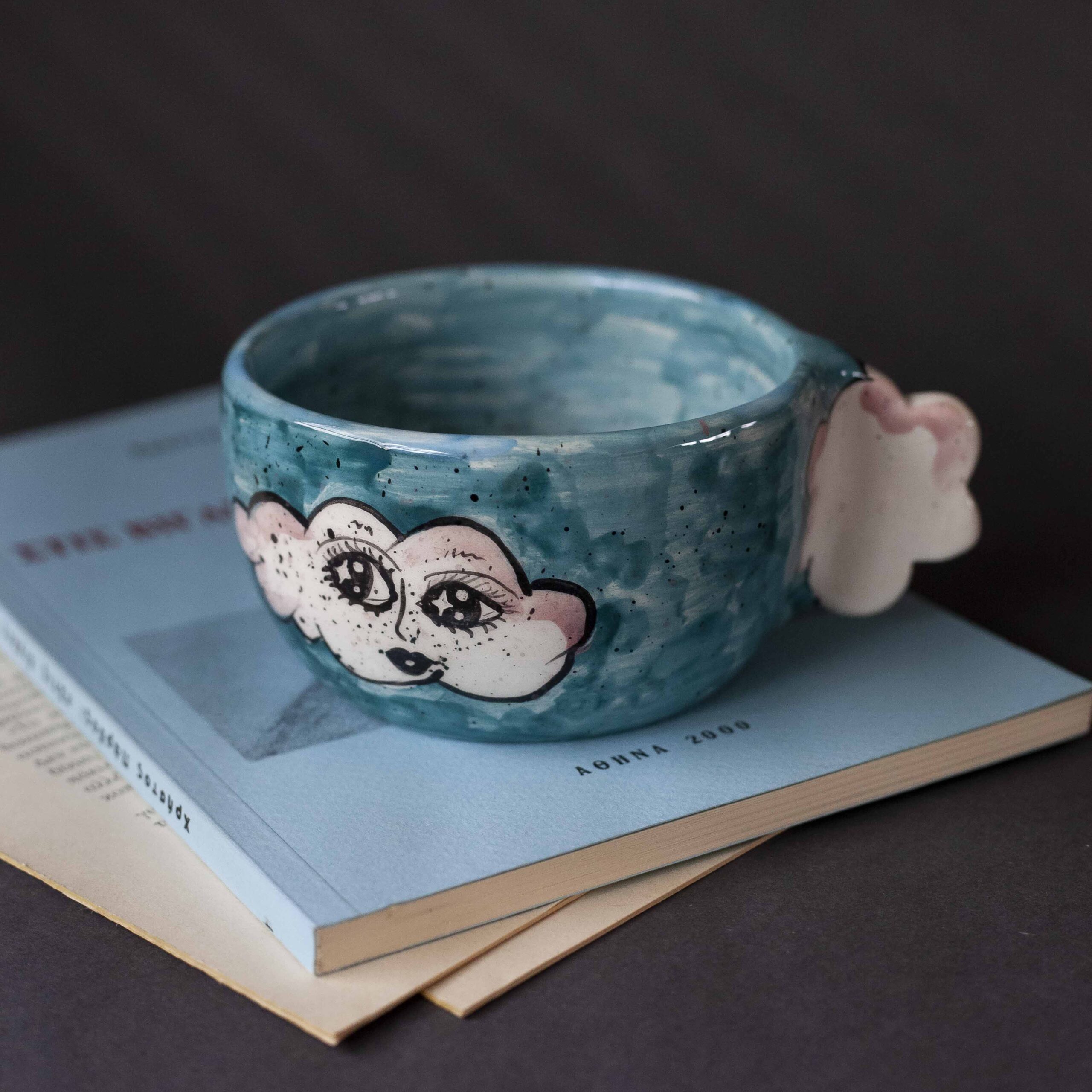 Handmade ceramic mug with a cloud-shaped handle and painted cloud faces featuring shiny eyes in an angelic core aesthetic. Soft blue and pink tones create a dreamy, sweet atmosphere. Each mug is one of a kind — hand-shaped and hand-painted, a small personal piece of art for those who love unique creations. Χειροποίητη κεραμική κούπα με χερούλι σε σχήμα συννεφιού και ζωγραφιστά συννεφάκια με εκφραστικά προσωπάκια και γυαλιστερά μάτια σε angelic core αισθητική. Οι απαλοί τόνοι του γαλάζιου και του ροζ προσδίδουν μια ονειρική, γλυκιά αίσθηση. Κάθε κούπα είναι μοναδική, πλασμένη και ζωγραφισμένη στο χέρι, ένα μικρό προσωπικό έργο τέχνης για κάθε λάτρη των ιδιαίτερων αντικειμένων.