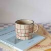 Medium handmade ceramic mug with pink checkerboard pattern, beige speckled stoneware, modern aesthetic Μεσαία χειροποίητη κεραμική κούπα με ροζ σκακιέρα, μπεζ πηλός με σαμότ, μοντέρνο σχέδιο