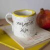 χειροποίητη κεραμική κούπα που γράφει χαμογέλα λιγο και από πισω έχει το χαμογελαστό ιμότζι. με κίτρινα χρώματα που δίνουν χαρά. handmade ceramic mug that writes smile a little . the yellow makes it more happy and it has a smiley imoji on behind.